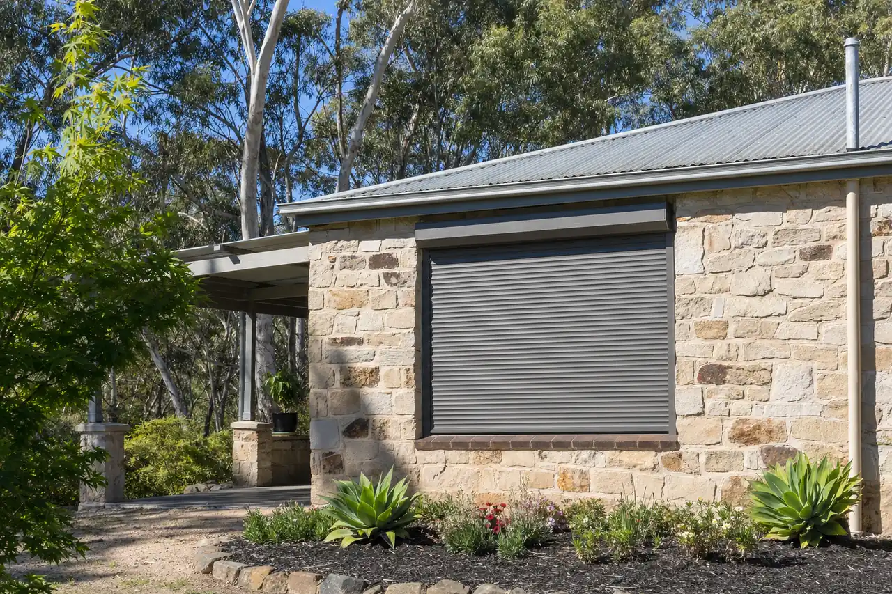 A closed metal shutter over a window on a stone house with eucalypt trees behind — bushfire shutters, Adelaide Hills. Illustrative example.